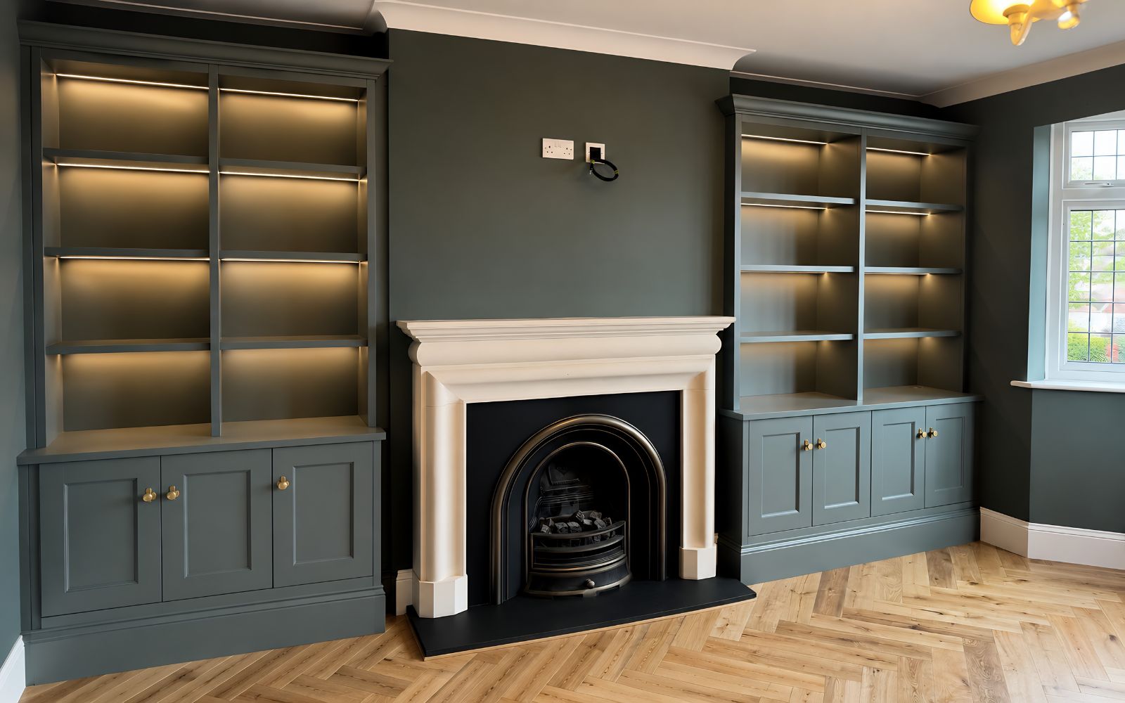 Bespoke grey-green alcove bookcases with LED-lit open shelves, shaker-style cabinets and brass knobs beside a traditional fireplace
