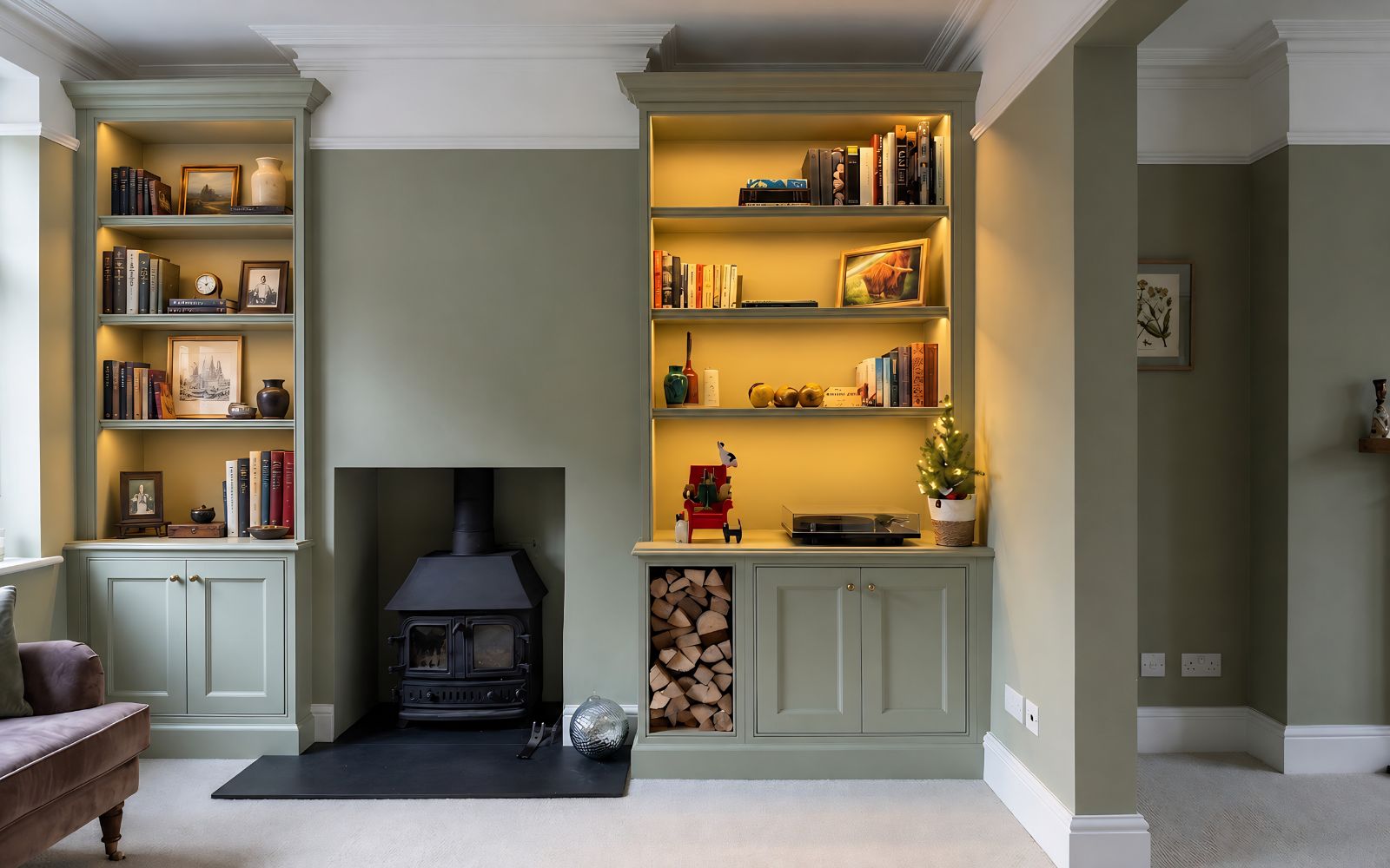 Bespoke sage-green alcove bookcases with LED-lit shelves, lower cabinets and log storage beside a wood-burning stove.