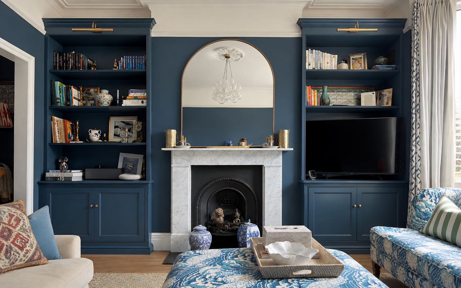 Bespoke blue alcove bookcases with lower cabinets, open shelves and TV recess beside a traditional marble fireplace.