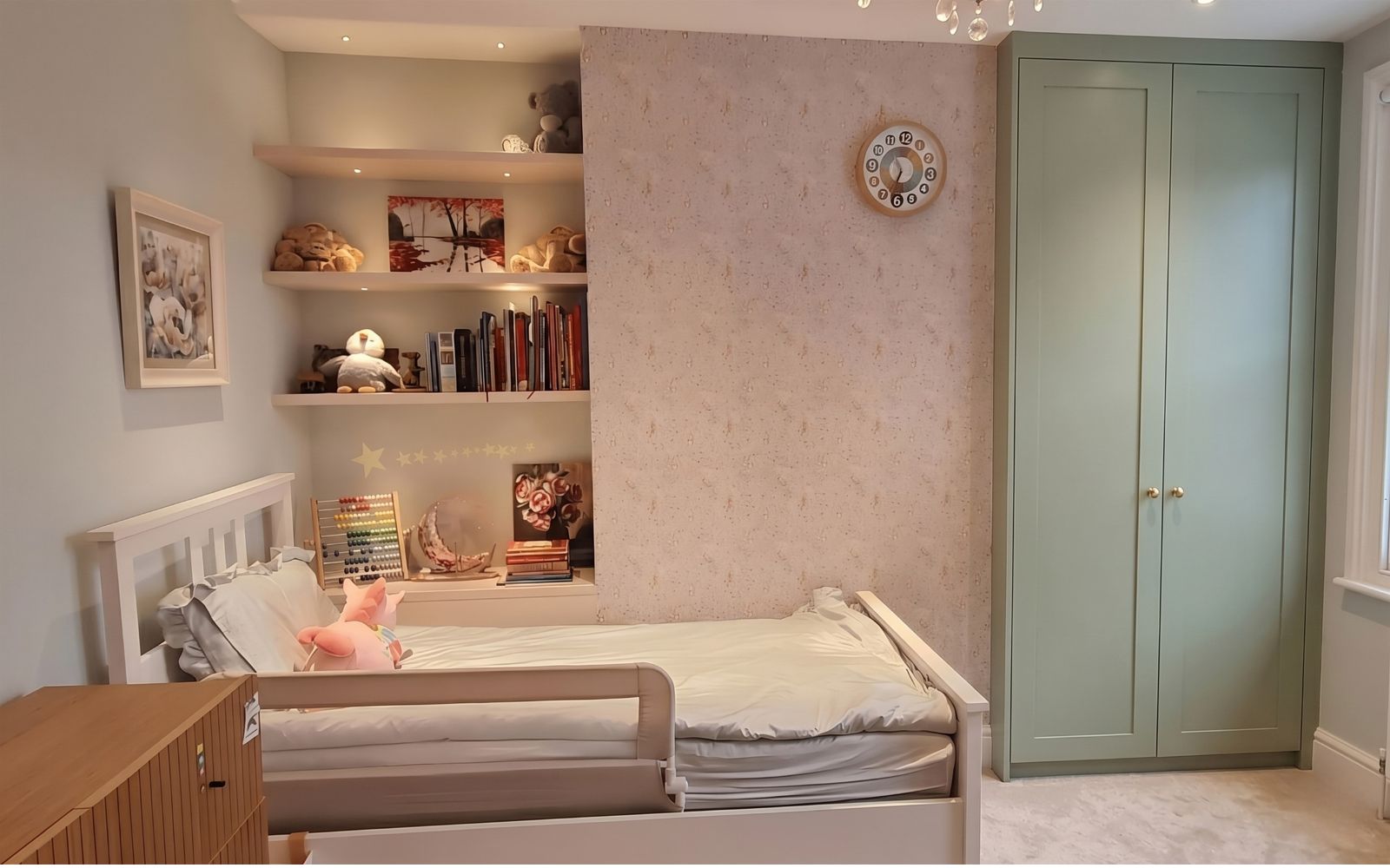 Children’s bedroom with bespoke open shelving, integrated lighting and a sage-green fitted wardrobe beside the bed.
