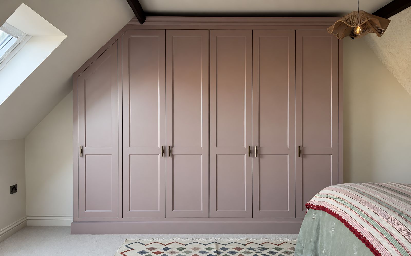 Bespoke dusky pink fitted wardrobe under a sloping loft ceiling, with shaker doors, brass handles, hanging rails and built-in drawers.
