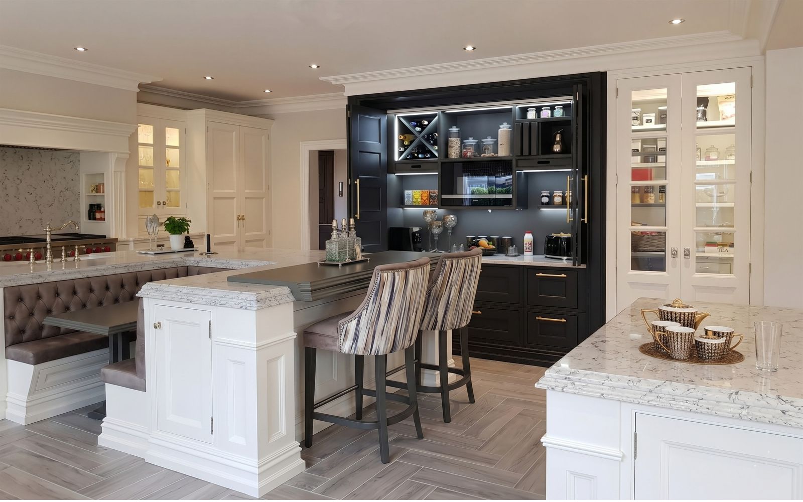 Bespoke luxury kitchen with painted cabinets, marble-effect worktops, black breakfast cupboard, island seating and glazed pantry storage.